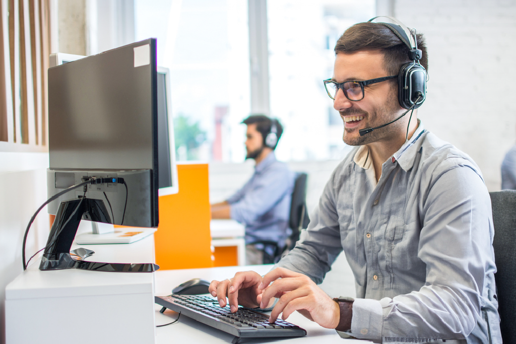 A man wearing a headset smiles and speaks while using a desktop computer in an office.