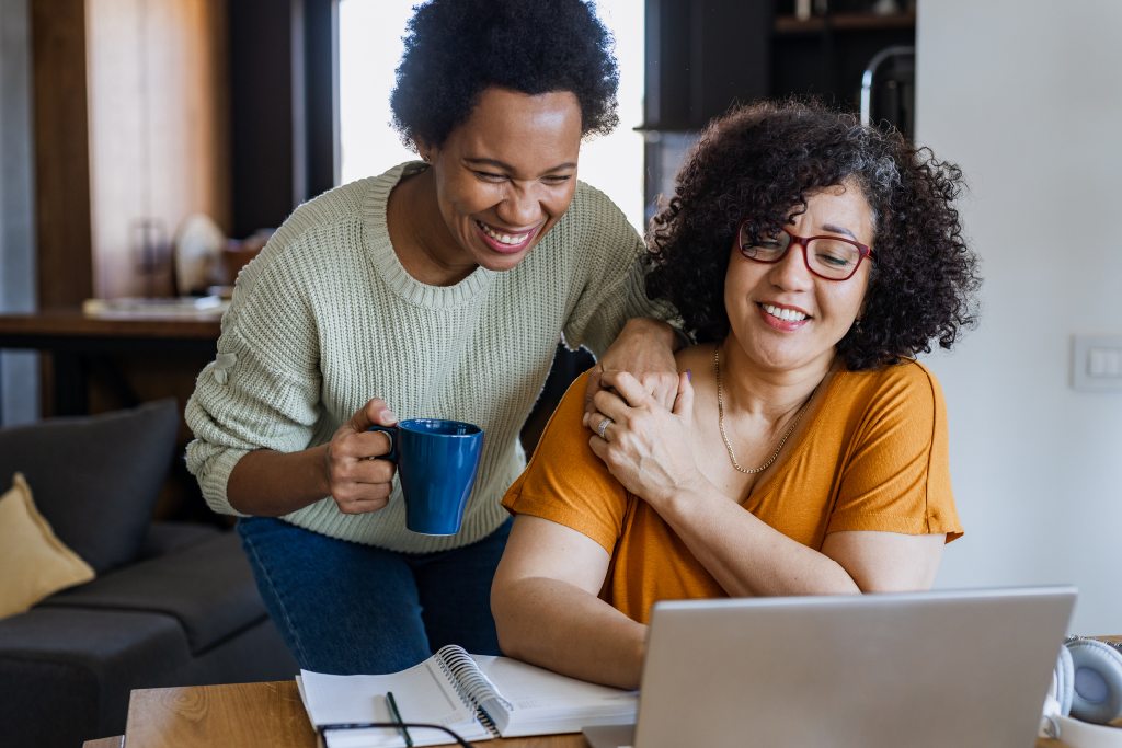 Two women holding hands and smiling hopefully at a laptop screen.