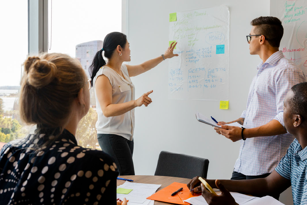 Four colleagues collaborate on a project at a whiteboard and table in an office.