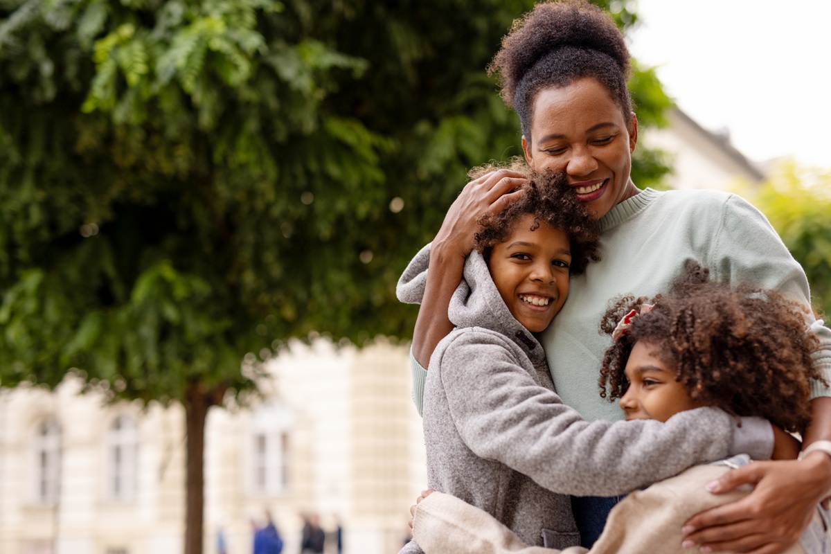 Mom with two children stand together hugging and smiling outside on a city street with tree.