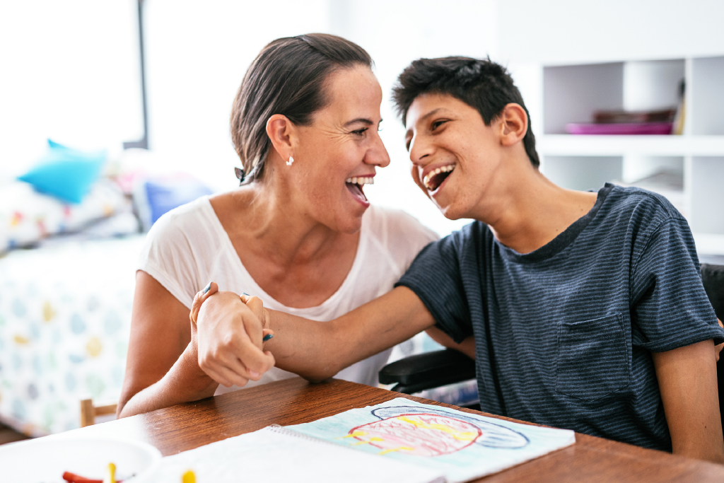 A mom and her son smiling at each other as they sit at a wooden table, with the mom holding his arm.