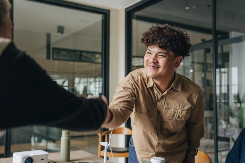 A young man smiling while shaking hands with another adult in an office.