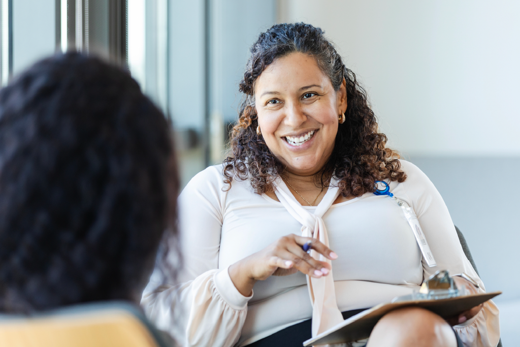A professionally dressed woman holding a clipboard and pen while smiling at someone across from her.